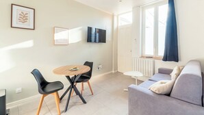 A modern living room with a grey sofa, a round wooden table with two black chairs, a flat-screen TV mounted on the wall, and a window with a blue curtain. - Charming T2 in the heart of Villeurbanne (Villeurbanne)