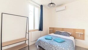 A bedroom with a bed, a wooden headboard, a window with curtains, a radiator, and a shelf unit. - Charming T2 in the heart of Villeurbanne (Villeurbanne)
