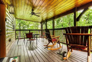 Relax on the cabin's spacious deck, surrounded by lush greenery, with a ceiling fan overhead and comfortable seating for two.
