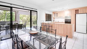 A modern kitchen with a central island, a dining area with a glass table, and a view of greenery outside. - Ocean Spray- Beachfront Retreat with Ocean Views, Jacuzzi & Pool (Yorkeys Knob)