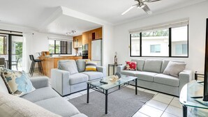 A modern living room with a sofa, coffee table, and a glass-top table. There is a kitchen area with wooden cabinets and a dining area with a bar. - Ocean Spray- Beachfront Retreat with Ocean Views, Jacuzzi & Pool (Yorkeys Knob)