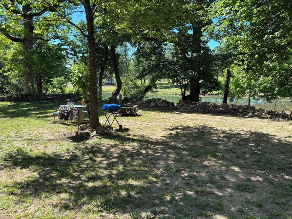 A grassy outdoor area with a barbecue grill, picnic table, and stone wall near a body of water.