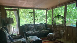 A cozy room with wicker furniture, a colorful striped rug, and large windows offering a view of greenery outside.