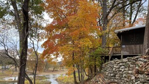 A rustic cabin with a stone wall and a wooden deck, surrounded by autumn foliage and a small pond.