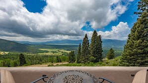 A panoramic view from a balcony with a metal table in the foreground, overlooking a valley with mountains and dense forests under a partly cloudy sky. - Spacious 4 bedroom with unmatched view! (Angel Fire)