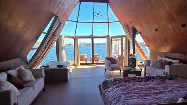 Una habitación amplia con techos altos, una cama grande, dos sofás, una mesa de centro y un televisor. Un ventanal ofrece vista al agua.