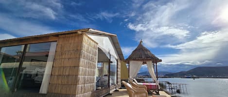 Una estructura de madera con amplios ventanales, un quiosco de techo de paja y una terraza de madera con sillas y una mesa.