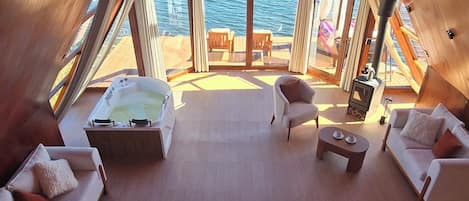 A high-ceilinged room with a large window overlooking water, furnished with a bed, sofas, and a small table.