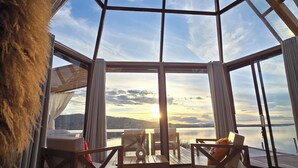Una habitación con un amplio ventanal de vidrio, muebles de madera y vista al atardecer sobre el agua.