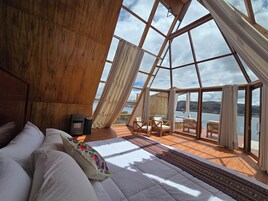 Una habitación con techo de madera, una cama con ropa de cama blanca, una pequeña mesa y vistas a un lago a través de grandes ventanales.
