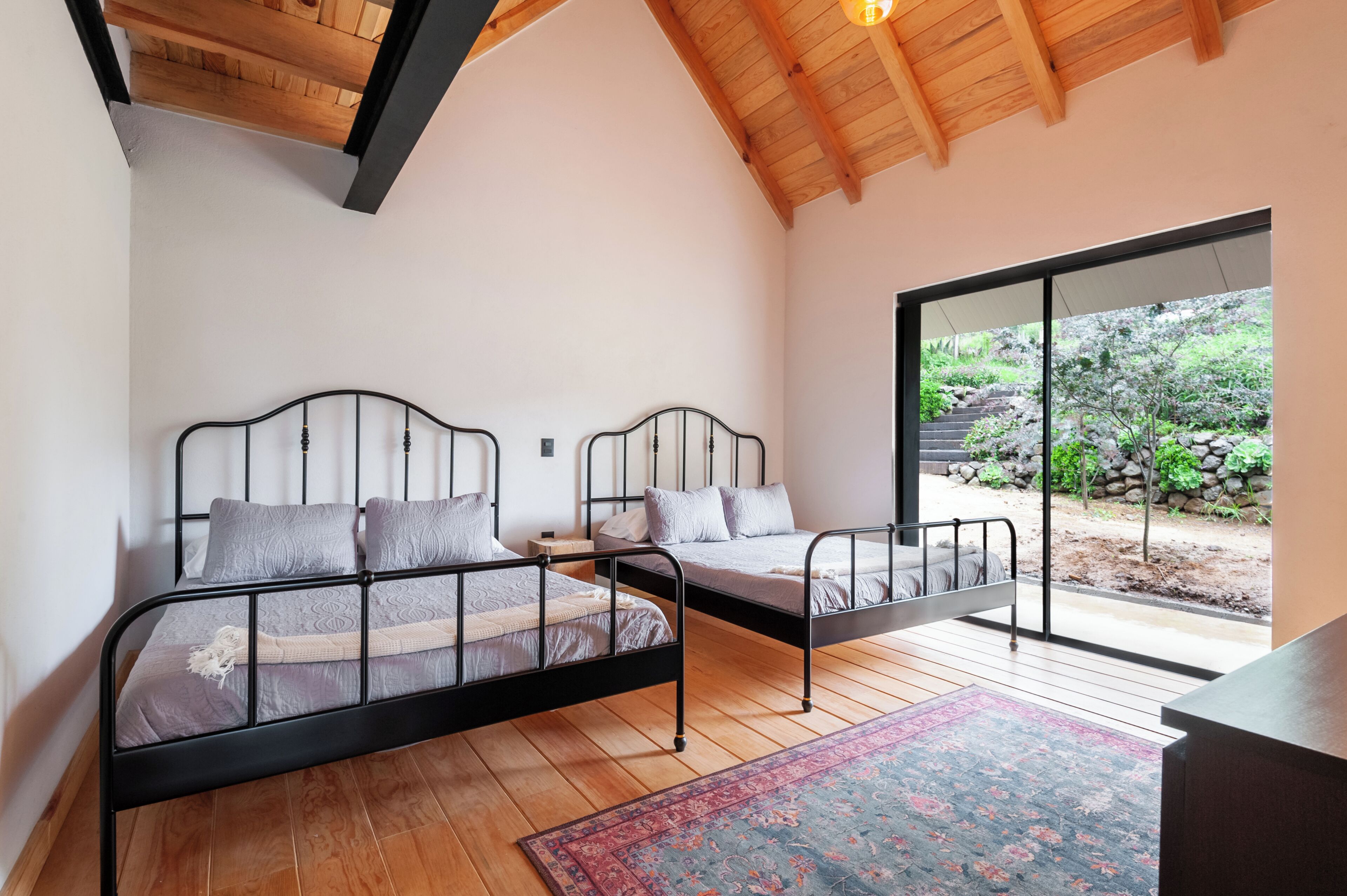 A bedroom with two beds, wooden flooring, a large window, and a view of a garden.