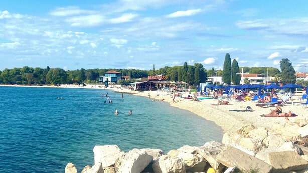 A beach with clear blue water, rocky shoreline, and a row of beach umbrellas. - Modern apartment in a perfect location. Sea and old town within walking distance (Fazana)