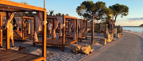 A beachside resort with wooden cabanas, a pebble beach, and a paved walkway.