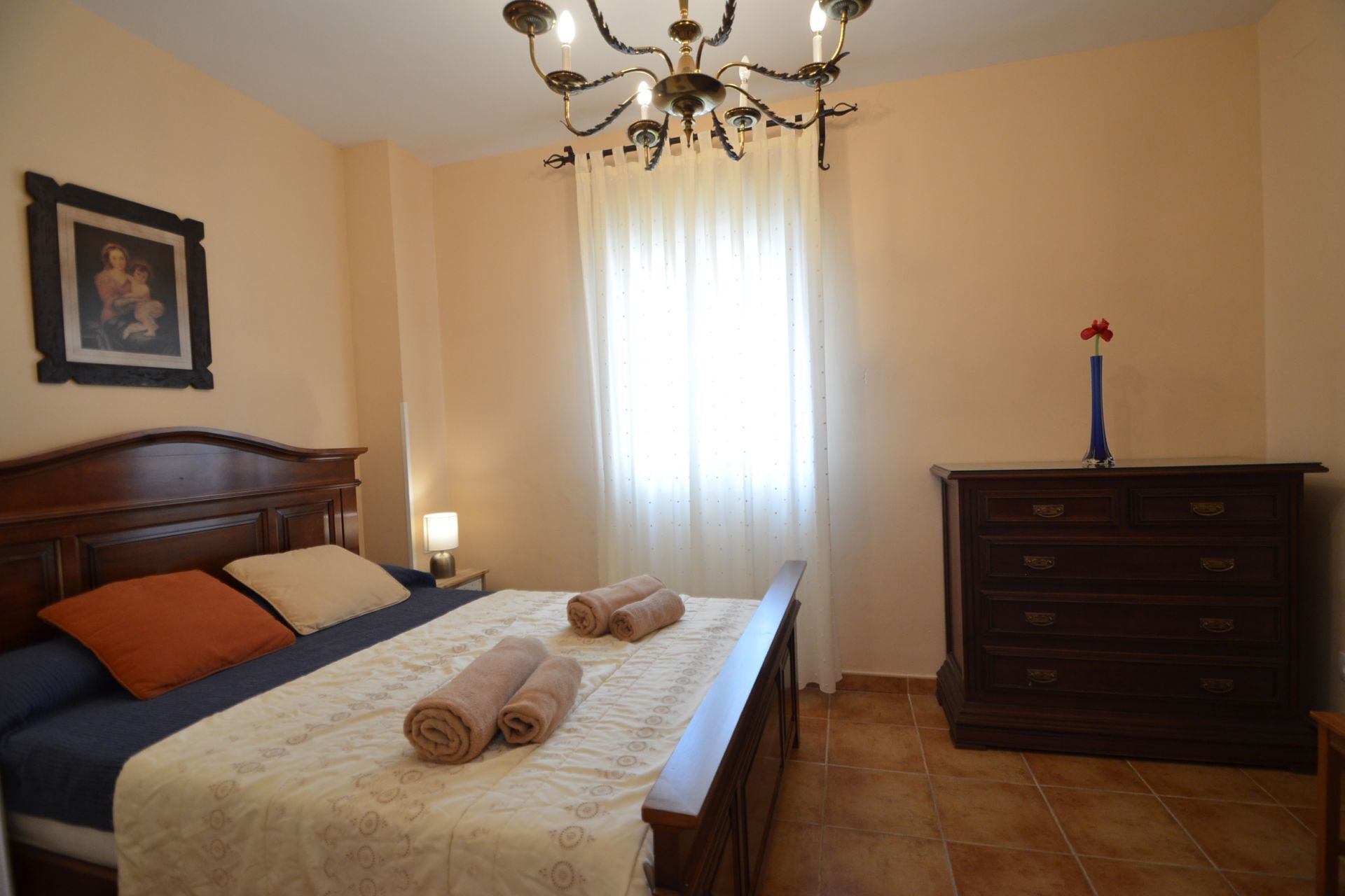 A bedroom with a bed, a nightstand, a dresser, a chandelier, and a window with curtains.