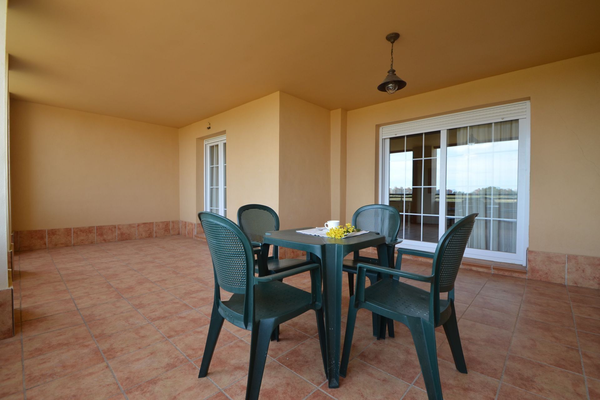 Une terrasse avec une table et quatre chaises, un ventilateur de plafond et une vue dégagée sur l’extérieur grâce à une grande fenêtre.