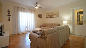 A living room with a sofa, a table, a ceiling fan, a clock, and a door.