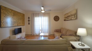 A room with a bed, a sofa, a small table, a lamp, a TV, and artwork on the walls.