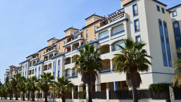 Un hôtel à plusieurs étages, doté de balcons, de palmiers et surplombant un ciel d’un bleu limpide.