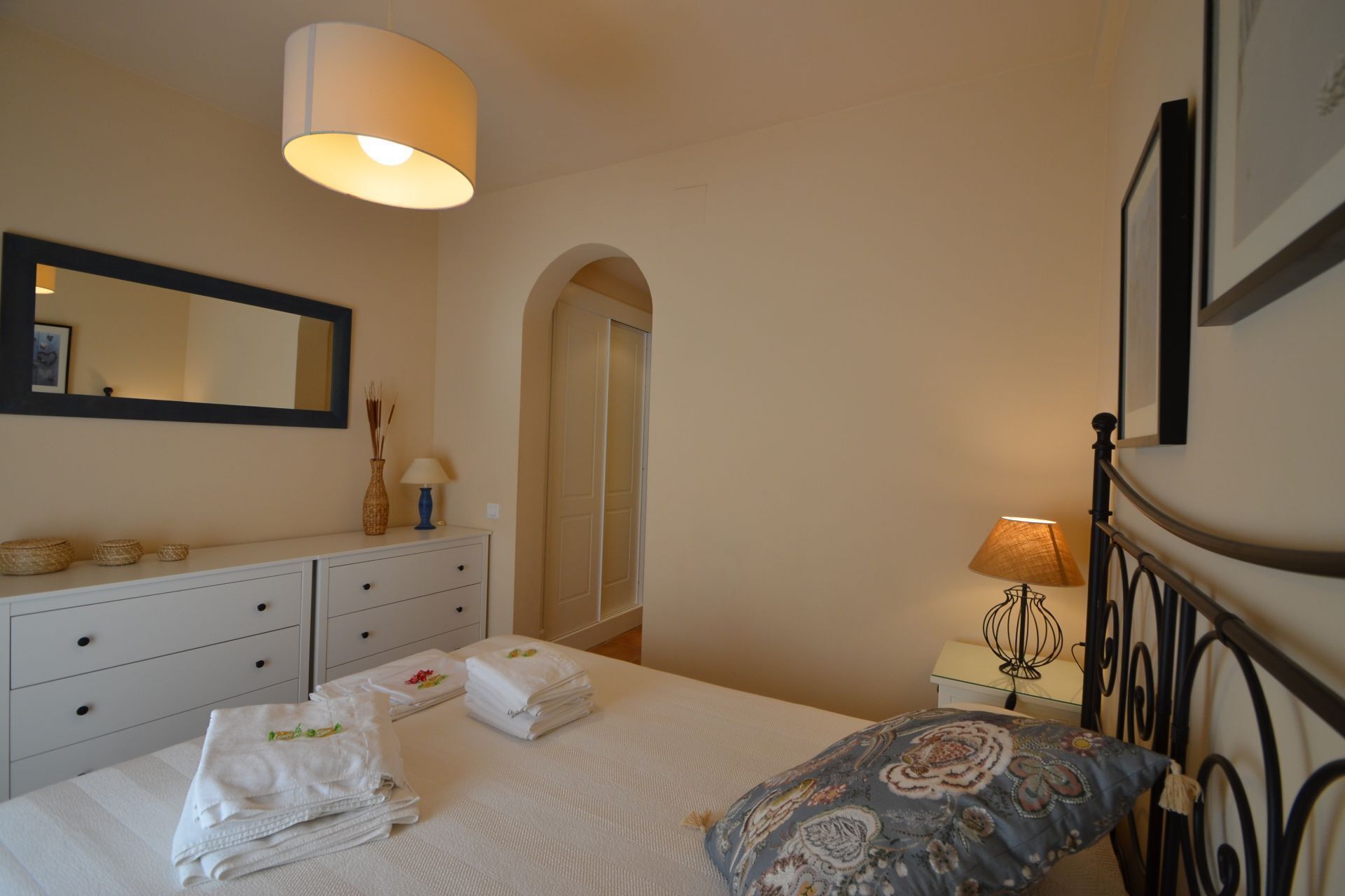 A hotel room with a bed, a nightstand with a lamp, a dresser, a mirror, and a door leading to another room.
