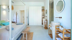 A modern bedroom with a bed, bench, and a bathroom with a stone basin and shelves. - Stunning Seafront Villa in the Mondragó National Park, Amazing Views & Direct Access to the Beach! (Cala Mondrago)