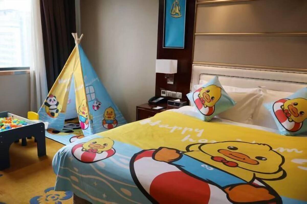 Habitación de hotel con cama y ropa de cama con temática de patos, un pequeño espacio de juegos con juguetes y una ventana con cortinas.