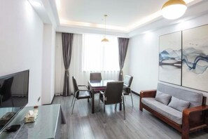 A hotel room with a TV, a dining table with chairs, a sofa, and a large window with curtains.