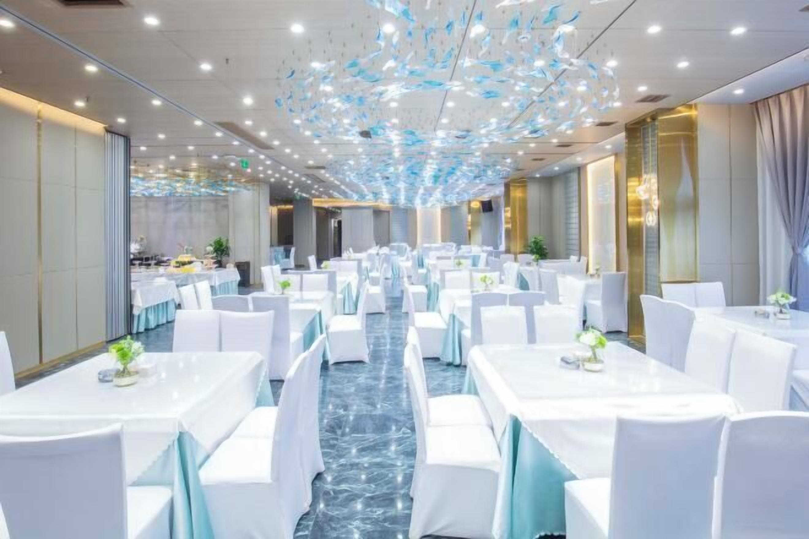 Une salle de banquet avec des tables et des chaises blanches, un sol en marbre et un plafond agrémenté d’un luminaire.