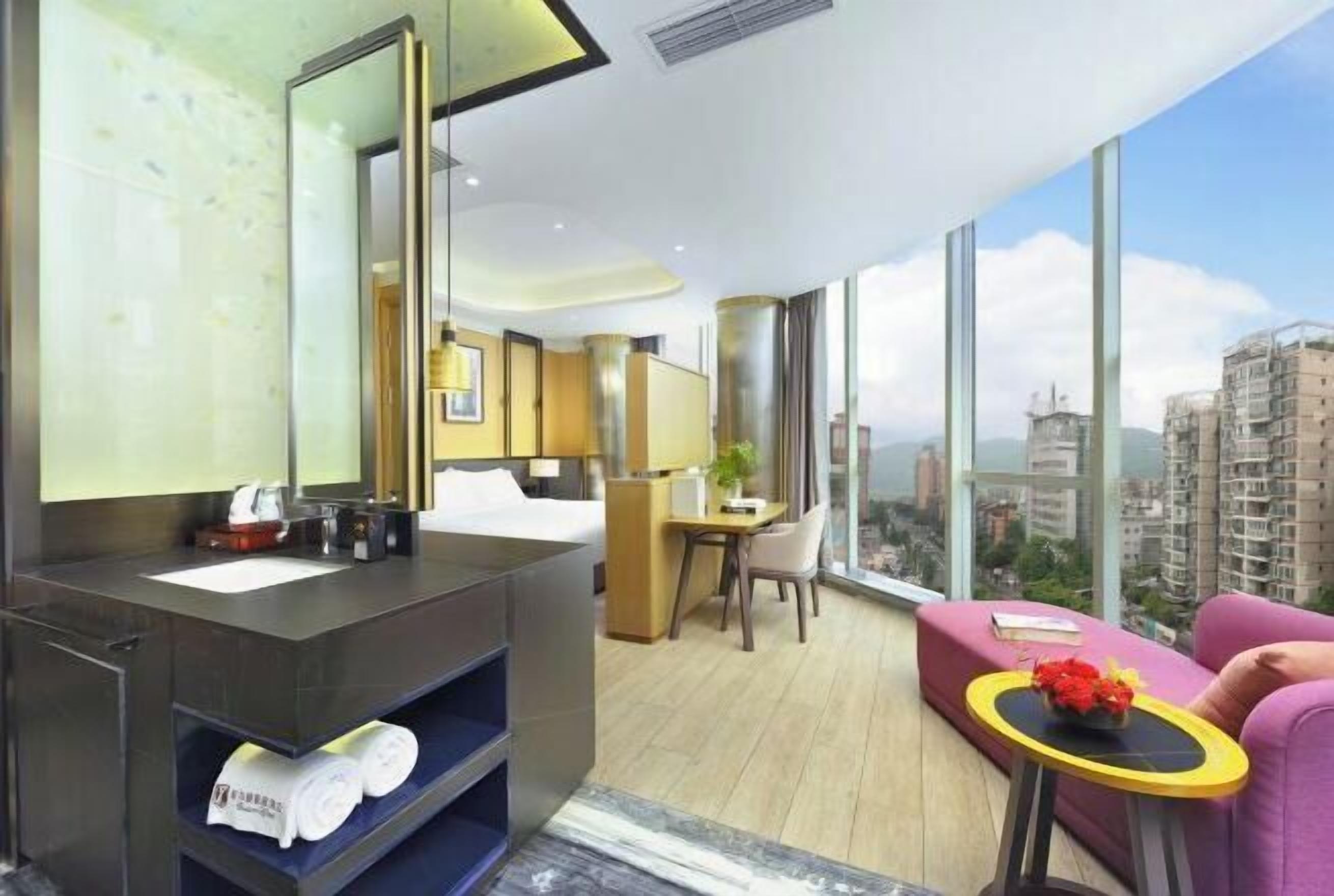 A modern hotel room with a large bed, a desk, a chair, a small table with flowers, and a balcony with a city view.