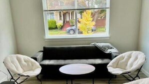 Une pièce avec un canapé noir, deux chaises blanches, une petite table ronde et une fenêtre avec des stores.