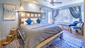 A bedroom with a bed, a ceiling fan, a window with curtains, a wooden bench, and a patterned rug. - The Rhododendron House is a spacious cottage in Waynesville, NC. It is ideal for families traveling with younger and older children and longer term stays. This open floor plan, 1000sq foot cottage is a stand alone building and has ample space to spre (Waynesville)