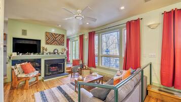 A cozy living room with a fireplace, a sofa, a television, and a large window with curtains.