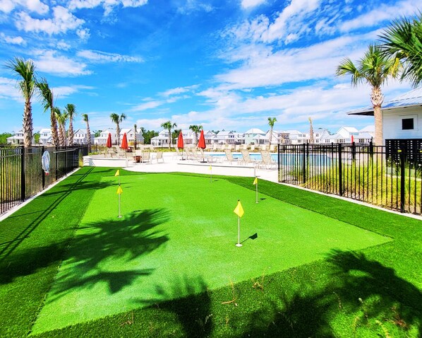 A golf course with a putting green, surrounded by a fence, palm trees, and a pool area with lounge chairs and umbrellas.