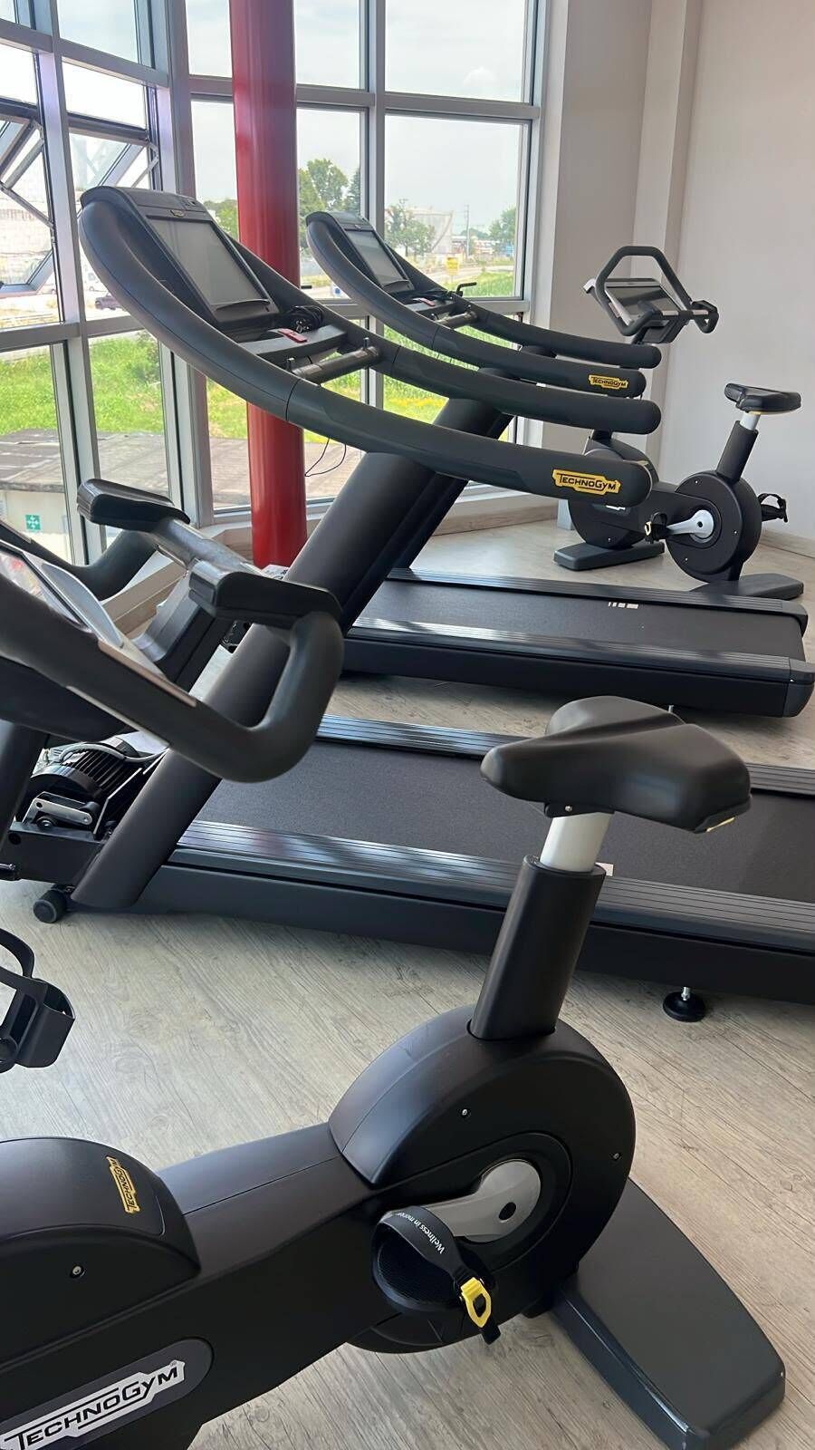 A gym with stationary bikes and treadmills, large windows, and a red pillar.