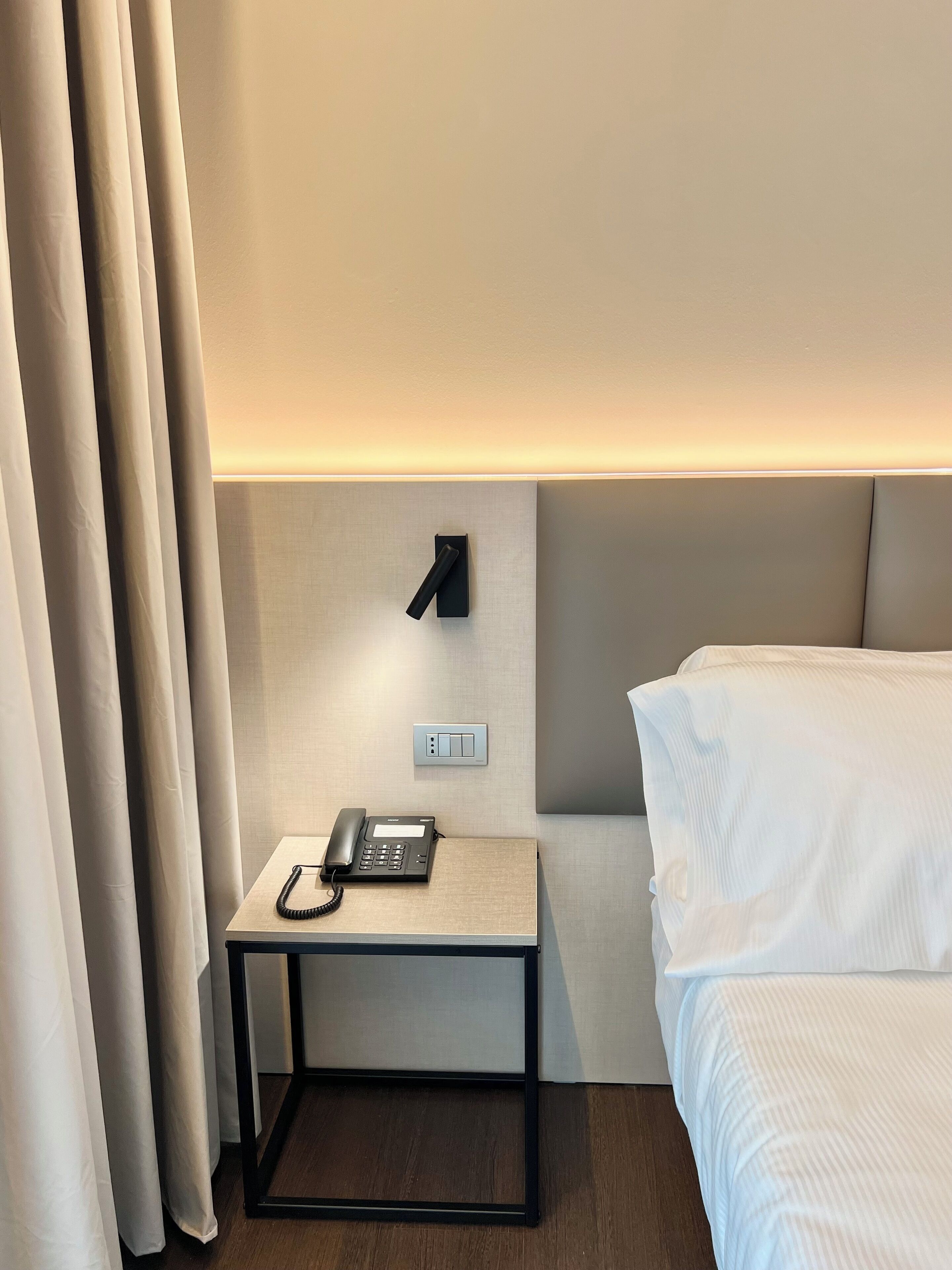Et hotelværelse med en seng, et natbord med en telefon og en lampe på væggen.