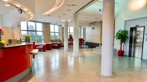 Un hall d’entrée spacieux avec un comptoir d’accueil rouge, des sièges confortables, de grandes fenêtres et un ascenseur moderne.