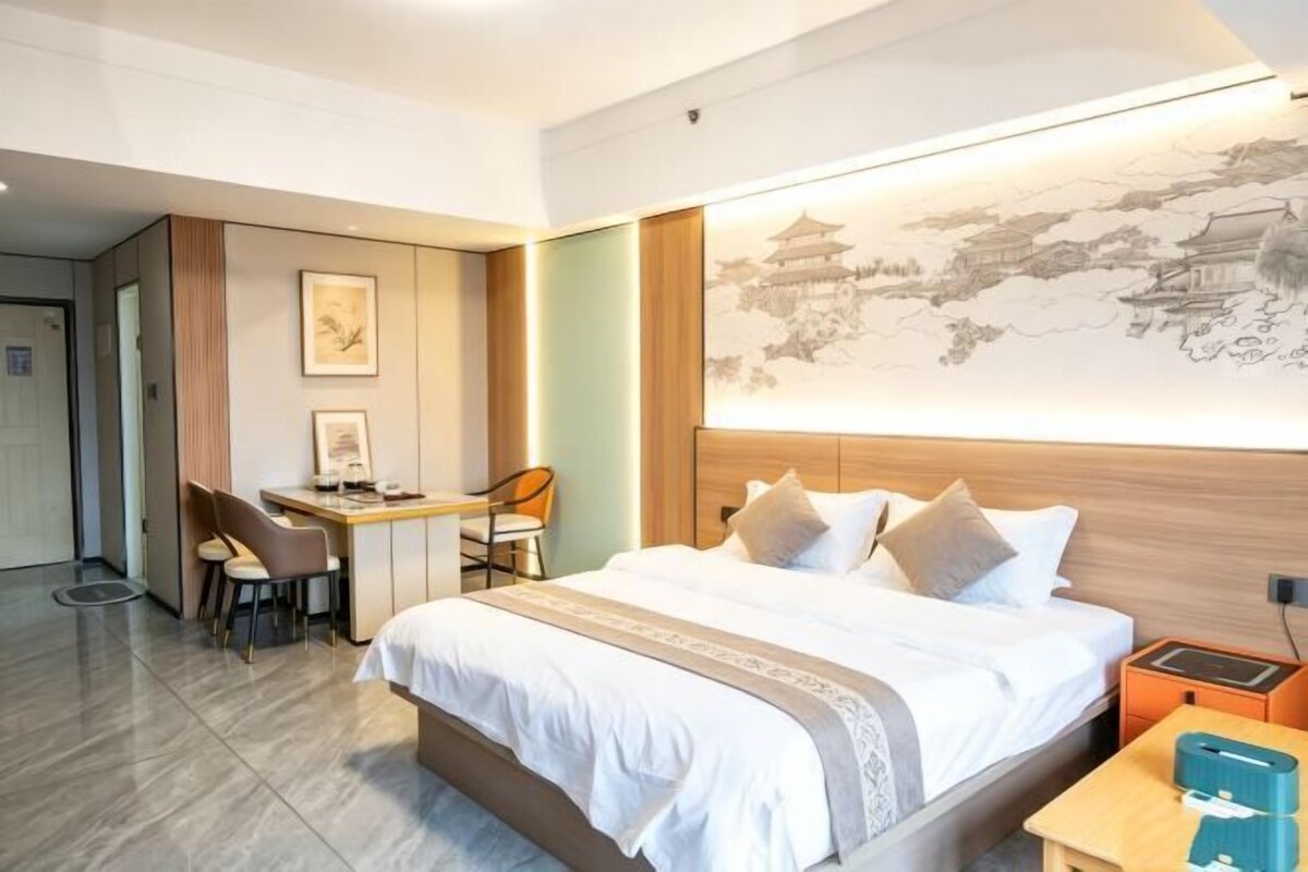 A hotel room with a bed, desk, and chair, a large artwork on the wall, and a traditional-style building in the background.