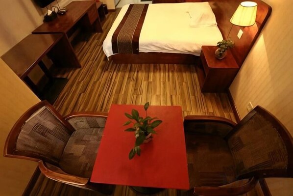 Habitación de hotel con una cama, una mesita con una planta, dos sillas, una mesita de noche con una lámpara y piso de madera.