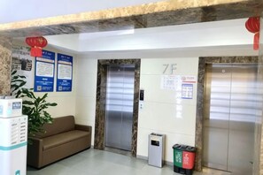 Un espace d’accueil comprenant deux ascenseurs, un canapé, une poubelle et des affiches d’information accrochées au mur.