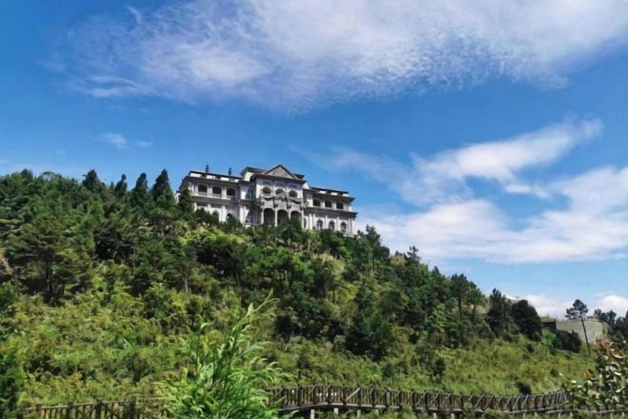 A large, historic building with multiple stories, surrounded by dense greenery and a clear sky.
