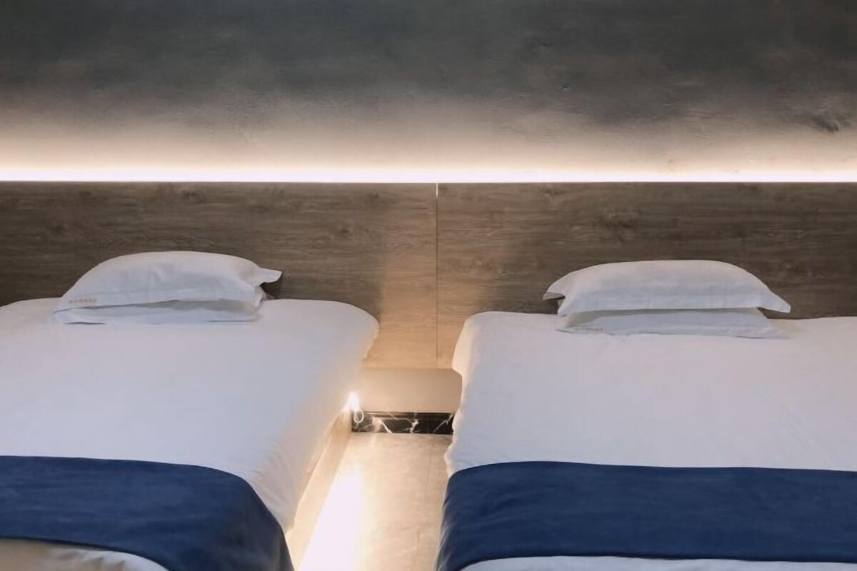 Two beds with white bedding and blue accents, a wooden headboard, and a small bedside table with a lamp.