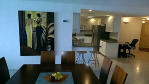 A dining area with a table set for four, a painting on the wall, and a kitchen in the background. - 4/4.5 Condo Oceanfront Cozumel Downtown AC WiFi Security (Cozumel)