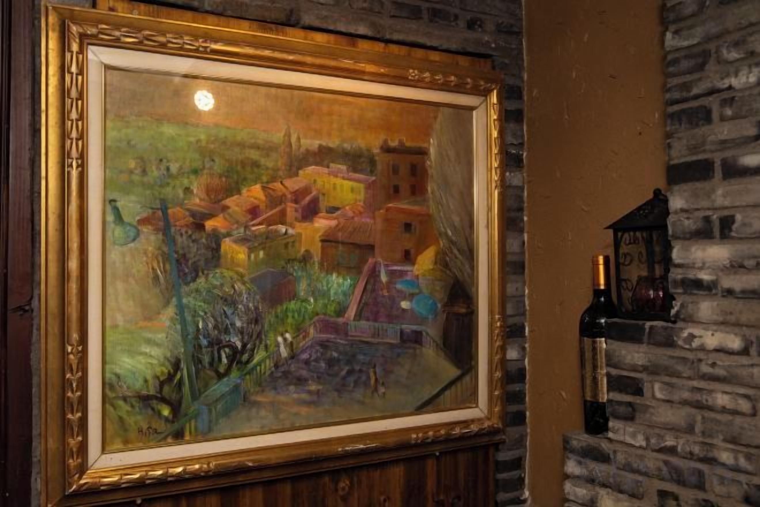 Un tableau encadré représentant un paysage de village, avec une bouteille de vin posée sur un mur de briques.