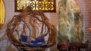 Een rieten stoel met een blauw kussen, een grote rots en een houten stoel voor een bakstenen muur met een lattenraam.
