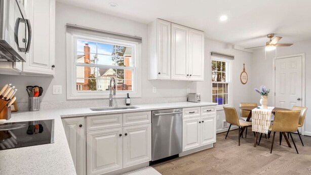A modern kitchen with white cabinetry, a stainless steel dishwasher, a window with a view of houses, and a dining area with a table and chairs. - Spacious 8 beds LUX convenient Home close to DC (Bethesda)