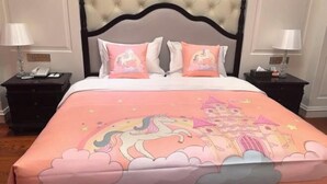 Une chambre à coucher avec une tête de lit capitonnée, un lit recouvert d’une couverture rose ornée d’une licorne et d’un château, deux tables de chevet avec des lampes et une petite table de nuit avec un téléphone.