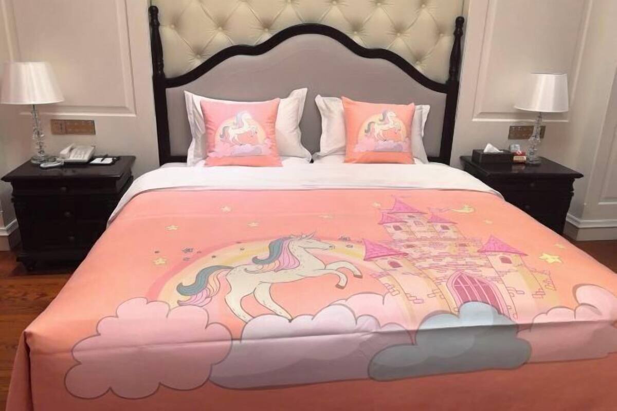 Une chambre à coucher avec une tête de lit capitonnée, un lit recouvert d’une couverture rose ornée d’une licorne et d’un château, deux tables de chevet avec des lampes et une petite table de nuit avec un téléphone.