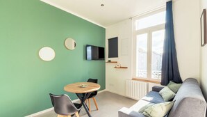 A modern living room with a green wall, a round wooden table with two chairs, a grey sofa with cushions, a flat-screen TV, and a window with curtains. - Cosy T2 completely renovated (Villeurbanne)