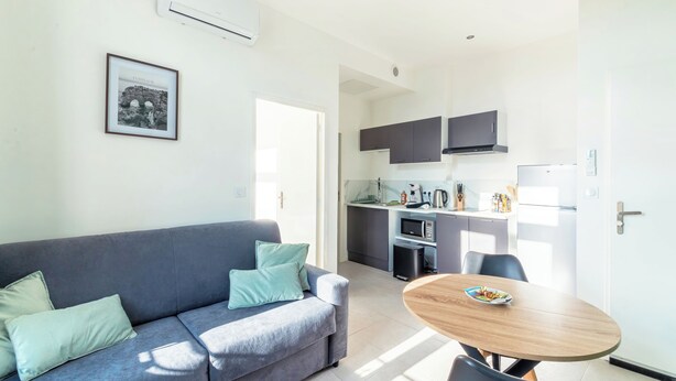 A modern living room with a grey sofa, a round wooden table, and a kitchen area with dark cabinets and a microwave. - Cosy T2 completely renovated (Villeurbanne)