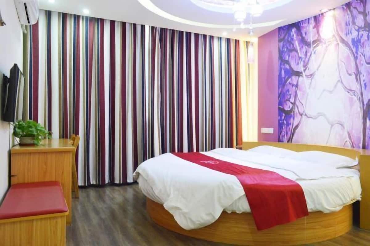 A hotel room with a round bed, a wooden nightstand, a red bench, a television, and a striped curtain.