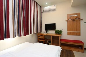 A hotel room with a bed, a desk with a chair, a TV, a plant, and a wardrobe with a mirror.